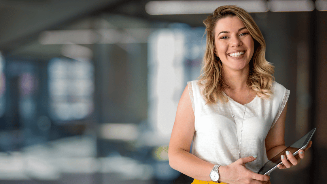 Smiling woman in a white sleeveless top holding a tablet in a modern office environment.
