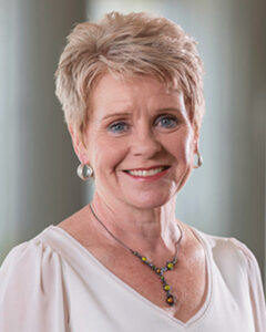 Smiling woman with short blonde hair wearing a white blouse and colorful beaded necklace.