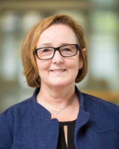 Smiling woman with short light brown hair and black glasses wearing a blue textured jacket indoors, blurred background