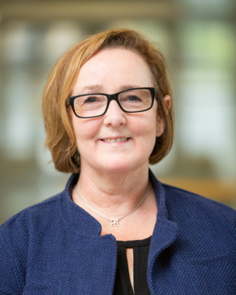 Smiling woman with short light brown hair and black glasses wearing a blue textured jacket indoors, blurred background
