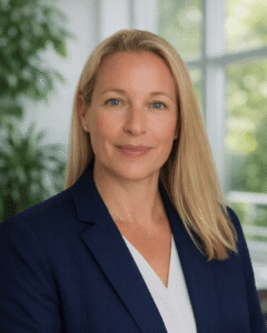 Professional headshot of a blonde woman in a navy blazer, smiling, in a bright office setting.