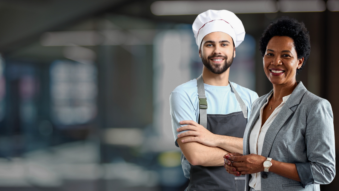 Smiling chef in a light blue shirt and gray apron stands with crossed arms beside a smiling woman in a gray blazer in a modern kitchen.