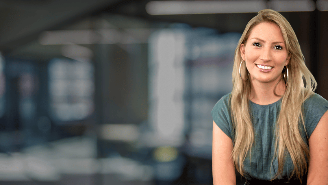 Smiling woman with long blonde hair in a blue-green top, in a blurred office background.