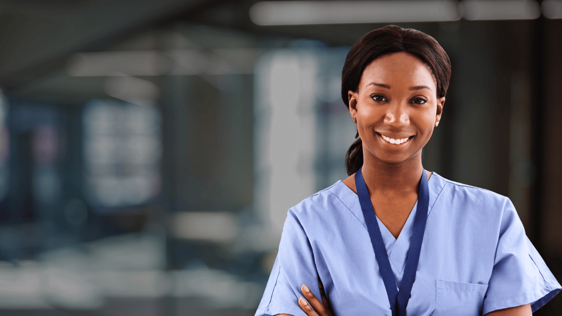 Smiling nurse in light blue scrubs with a blue lanyard standing in a hospital corridor, conveying a welcoming healthcare professional image.