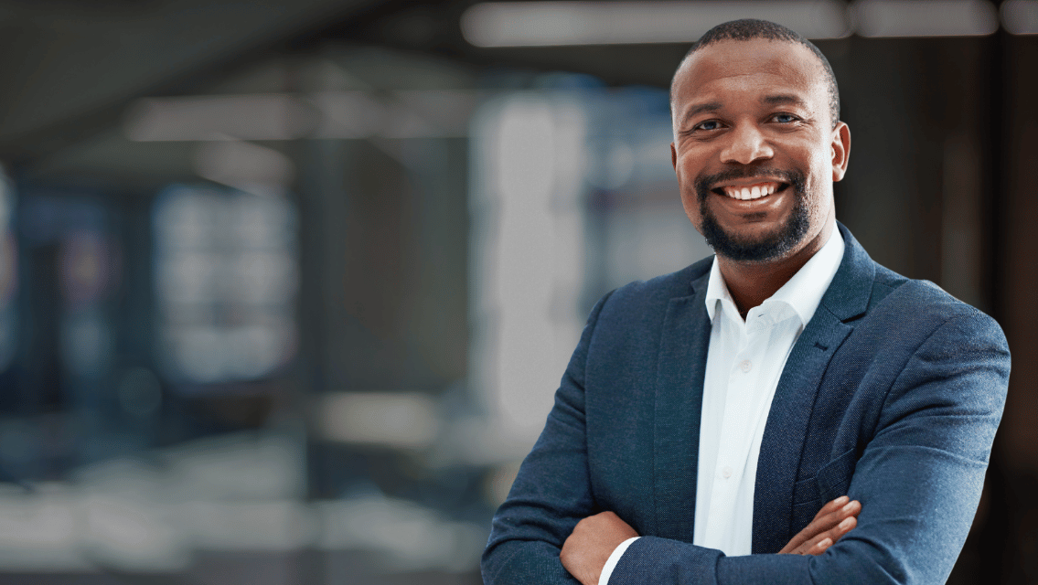 Smiling man in a navy blazer and white shirt with arms crossed in a modern office setting.