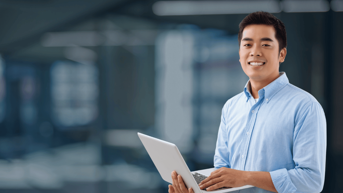 Smiling man in a light blue shirt holding a laptop in a modern office setting.