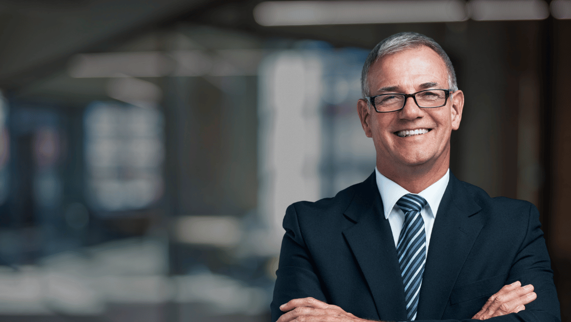 Confident businessman in a dark suit and striped tie, arms crossed, smiling in a modern office.