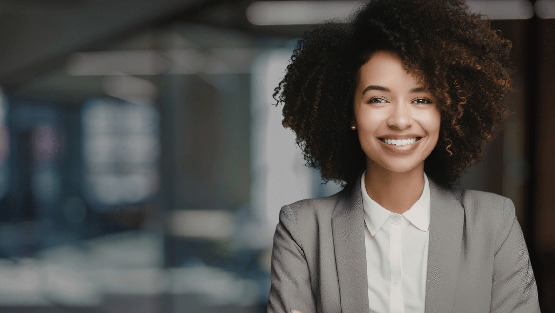 Professional woman with curly hair in a gray blazer and white shirt, smiling in an office setting with blurred background.