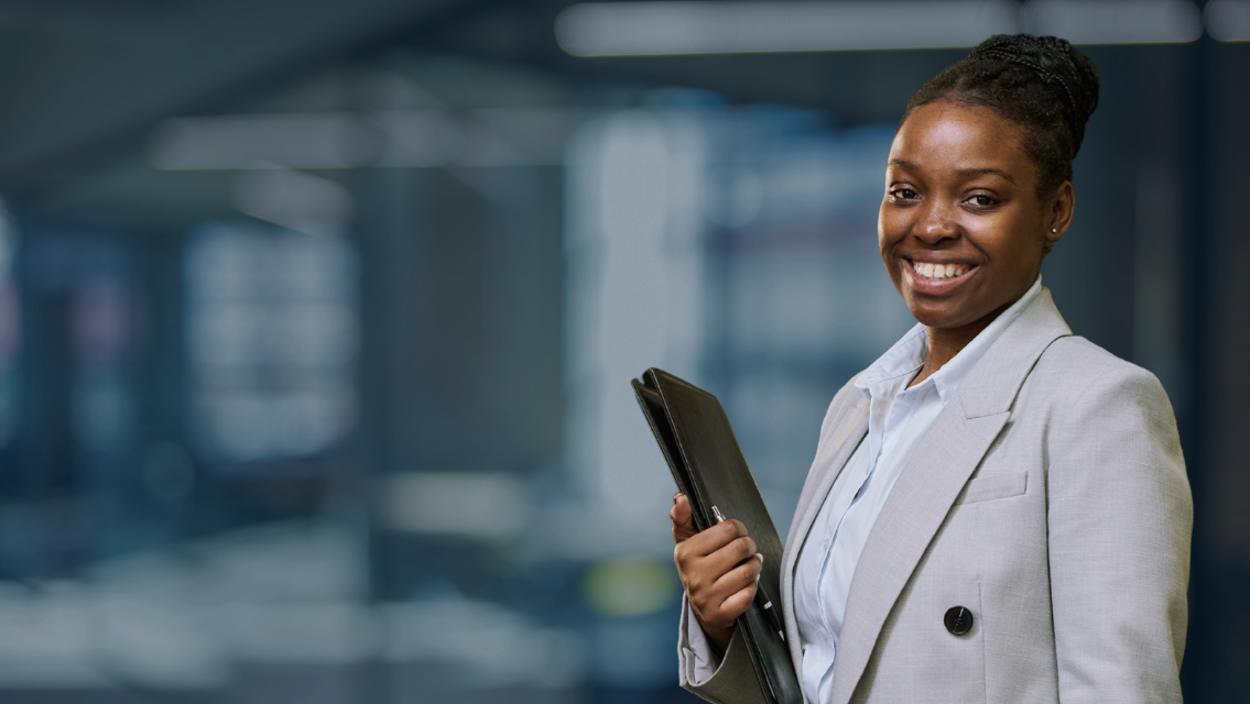 Smiling Black woman in a light gray blazer holds a tablet in a modern office, presenting a professional demeanor.